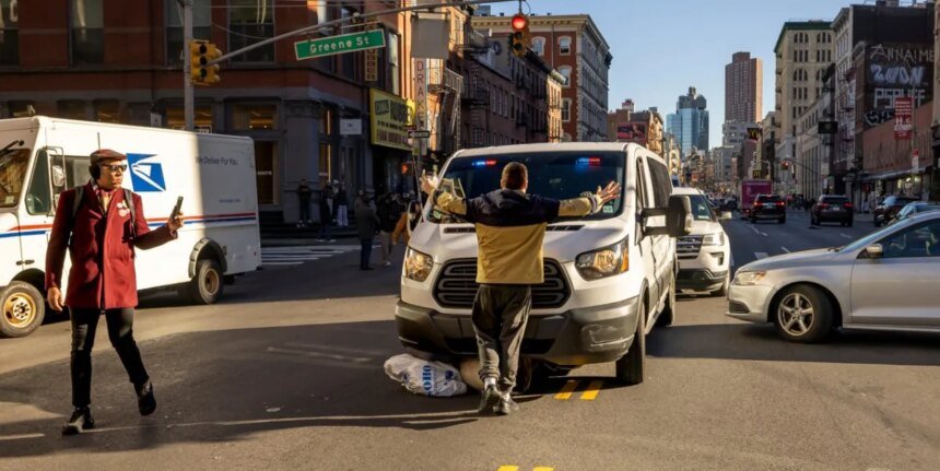 Man in a beige jacket stands with arms spread in front of a white delivery van stopped in the middle of a busy city intersection, with pedestrians and tall buildings around.