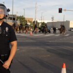Uniformed police officer in riot gear with helmet and face shield stands in the street, baton in hand, with officers on horseback in the background.