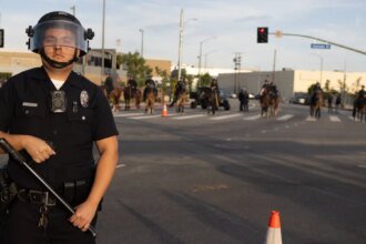 Uniformed police officer in riot gear with helmet and face shield stands in the street, baton in hand, with officers on horseback in the background.