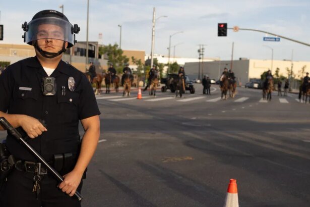 Uniformed police officer in riot gear with helmet and face shield stands in the street, baton in hand, with officers on horseback in the background.