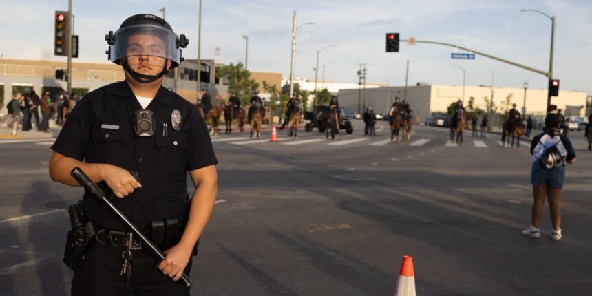 Uniformed police officer in riot gear with helmet and face shield stands in the street, baton in hand, with officers on horseback in the background.