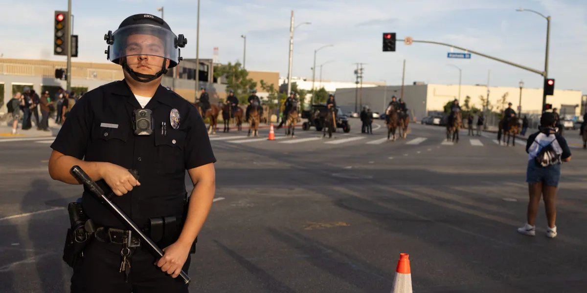 Uniformed police officer in riot gear with helmet and face shield stands in the street, baton in hand, with officers on horseback in the background.