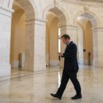 Businessman in a dark suit walks through a marble arched hall while looking at his phone.