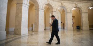 Businessman in a dark suit walks through a marble arched hall while looking at his phone.