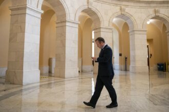 Businessman in a dark suit walks through a marble arched hall while looking at his phone.