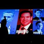 Silhouette of a person pointing at a large colorful wall of campaign portraits and polling percentages, featuring Rubio at center and other candidates like Harris, Carlson, Trump, Buttigieg, Ossoff, and Johnson nearby.