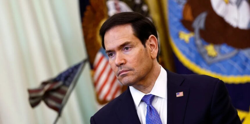 Man in a dark suit and blue tie with a small American flag pin, at a formal event with flags in the background, looking thoughtful or serious.