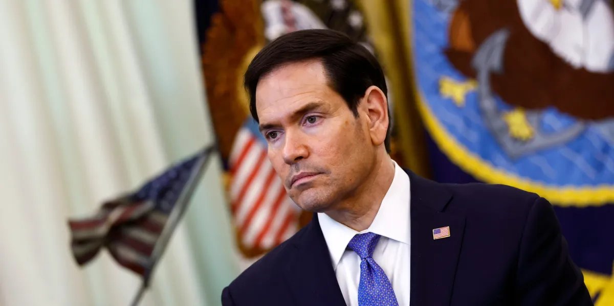 Man in a dark suit and blue tie with a small American flag pin, at a formal event with flags in the background, looking thoughtful or serious.