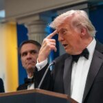 Man in a black tuxedo and bow tie speaks at a podium, with two suited men behind him and an American flag in the background.