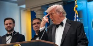 Man in a black tuxedo and bow tie speaks at a podium, with two suited men behind him and an American flag in the background.