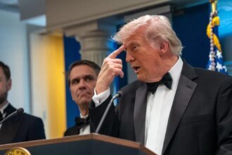 Man in a black tuxedo and bow tie speaks at a podium, with two suited men behind him and an American flag in the background.