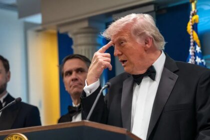 Man in a black tuxedo and bow tie speaks at a podium, with two suited men behind him and an American flag in the background.