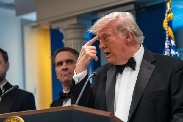 Man in a black tuxedo and bow tie speaks at a podium, with two suited men behind him and an American flag in the background.