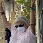 Woman wearing a face mask and sunglasses raises both arms in celebration outside a brick building, as bystanders film with their phones.
