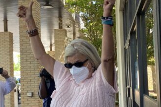 Woman wearing a face mask and sunglasses raises both arms in celebration outside a brick building, as bystanders film with their phones.