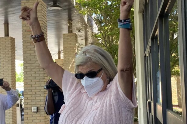 Woman wearing a face mask and sunglasses raises both arms in celebration outside a brick building, as bystanders film with their phones.