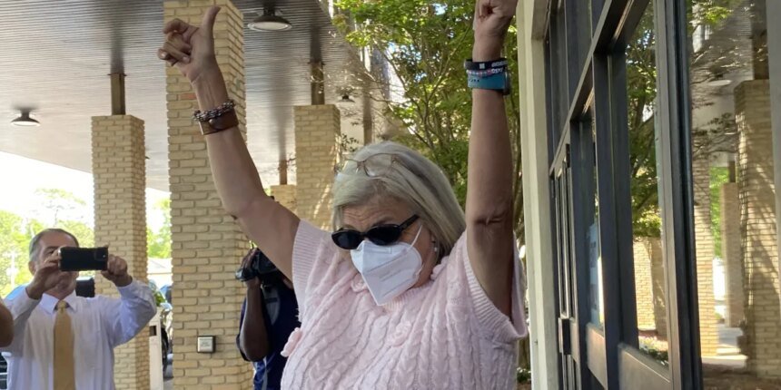 Woman wearing a face mask and sunglasses raises both arms in celebration outside a brick building, as bystanders film with their phones.